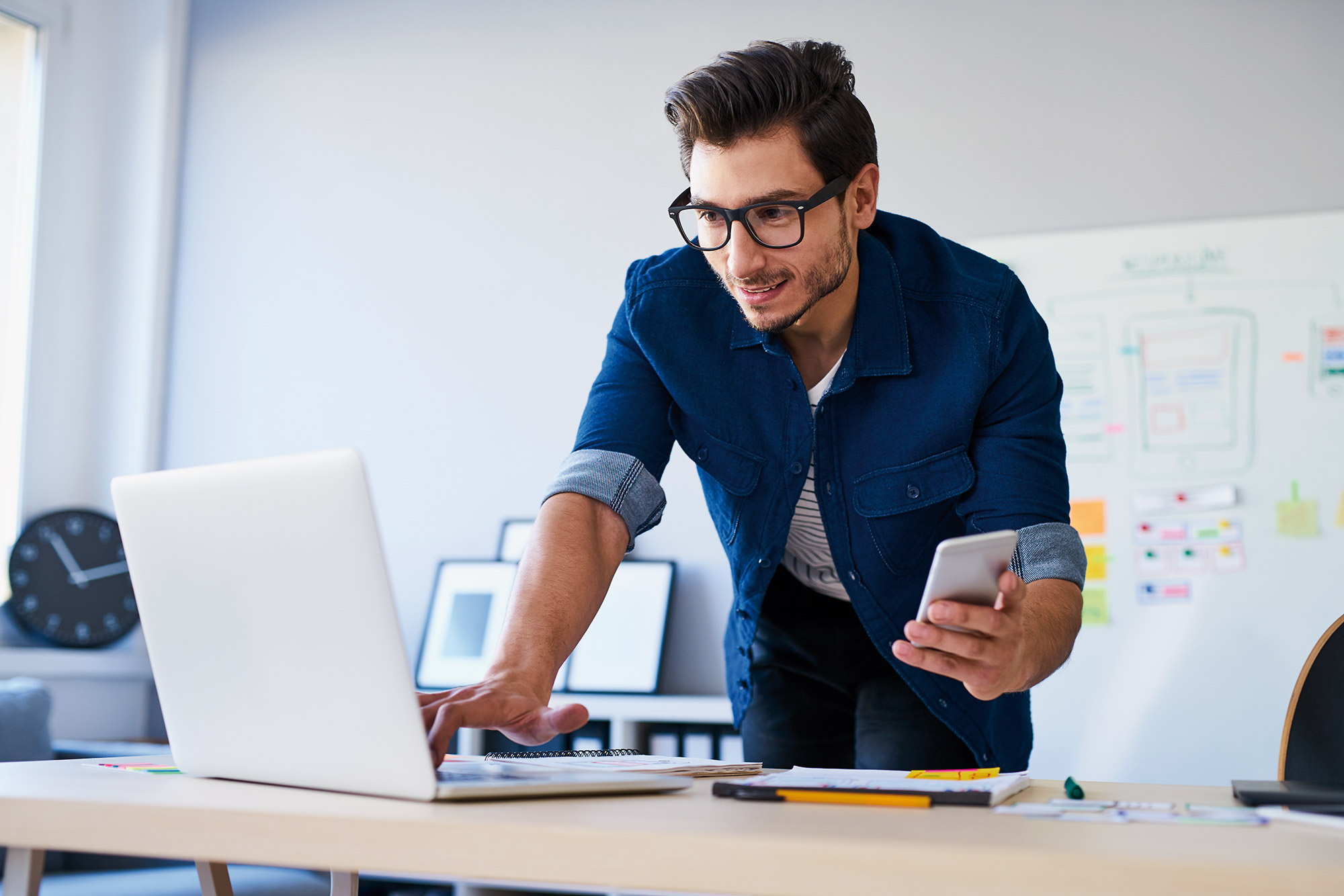 Ein Mann mit einem Smartphone in der Hand tippt etwas auf einem Laptop